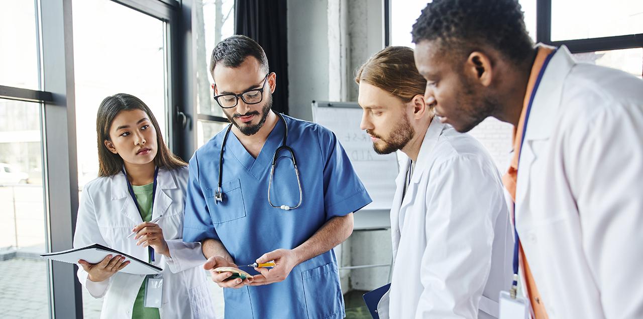 young male and female doctors in white lab coats taking notes and learning from older doctor in blue scrubs.